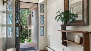 Un couloir doté d’un plancher en bois, une porte avec des panneaux de vitrail, une étagère fixée au mur sur laquelle se trouve une plante, et un miroir placé au-dessus.