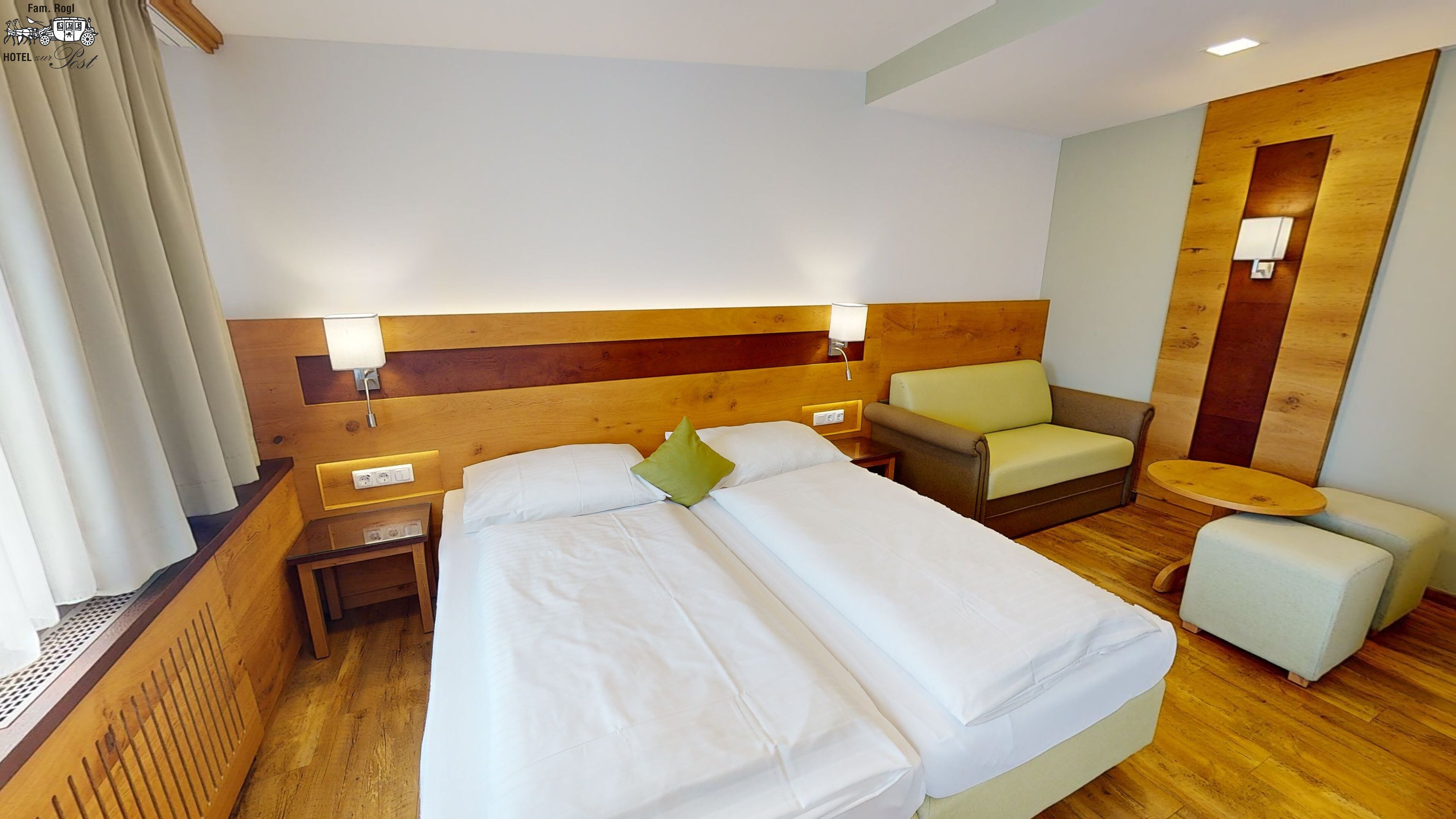 A hotel room with two beds, a wooden headboard, a small armchair, a round table, and a nightstand with a lamp.