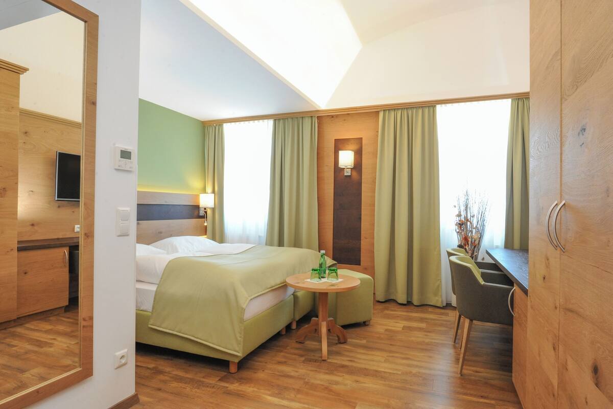 A hotel room with a bed, a small table with a bottle and glass, a chair, a desk, and a mirror.