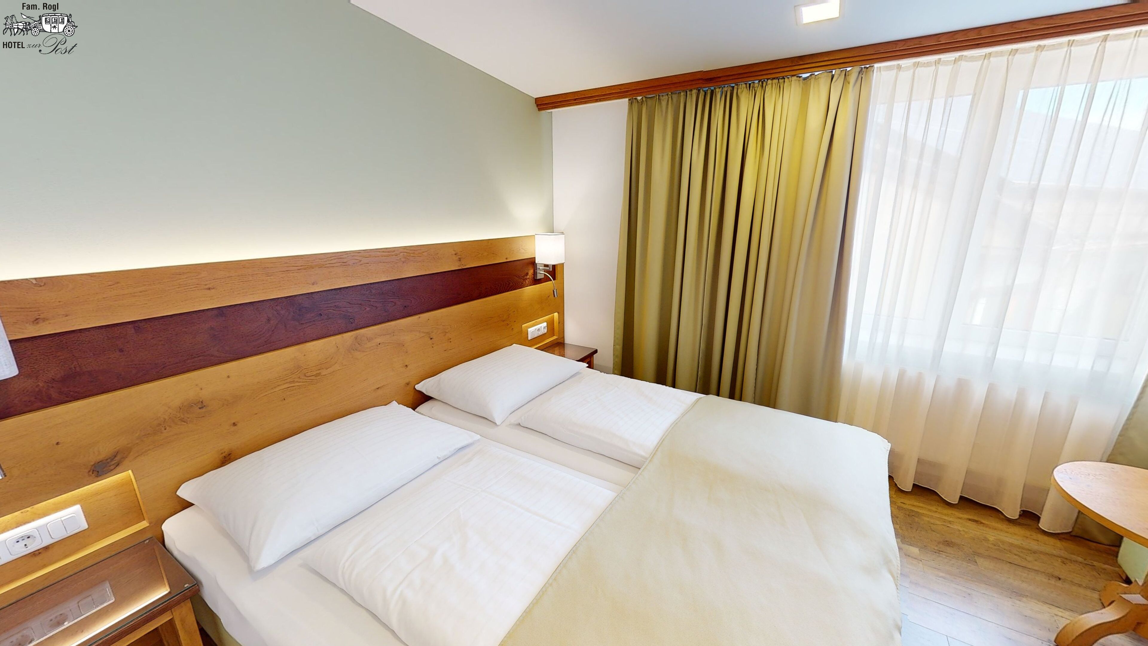 A hotel room with a wooden headboard, a bed with white linens, a bedside table, a lamp, and a window with curtains.