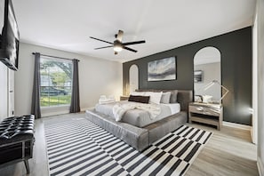 A bedroom with a bed, bedside tables, a ceiling fan, a window with curtains, and a view of the outdoors. - Backyard Resort w/ Pool, Spa, Sauna + Theater (Austin)