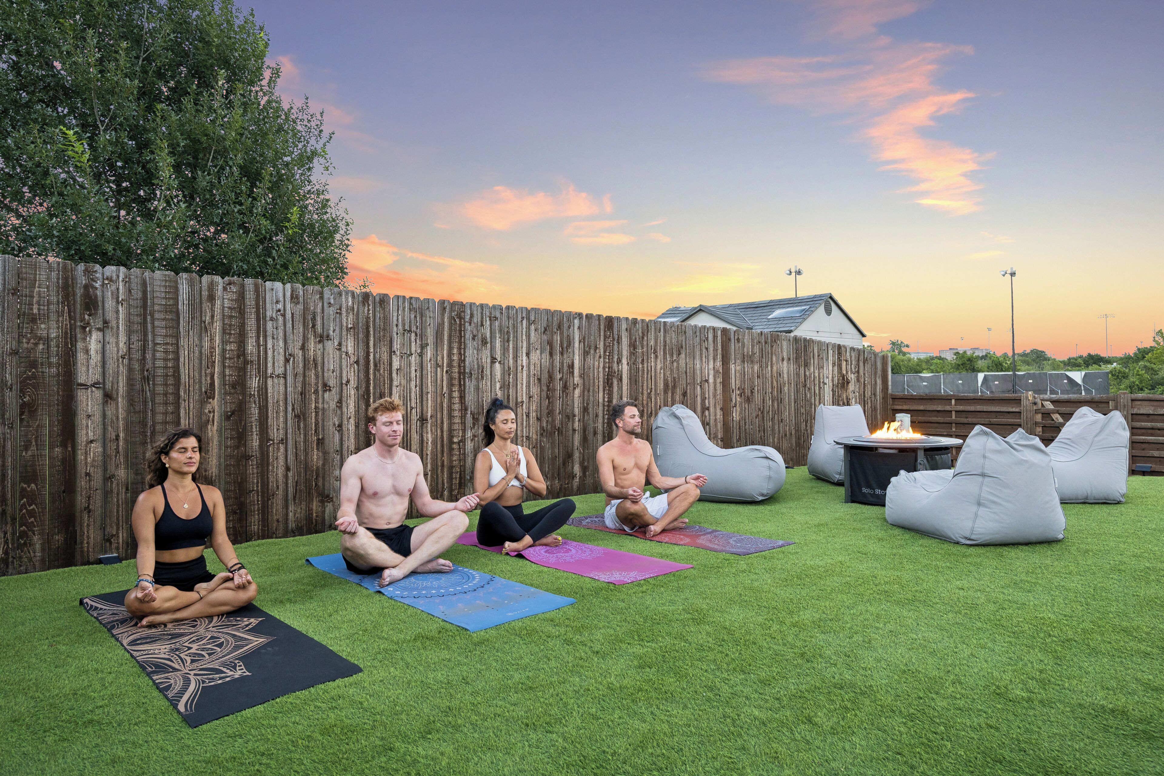 Un grupo de personas sobre esterillas de yoga en un espacio exterior con una valla de madera, césped y un atardecer de fondo.