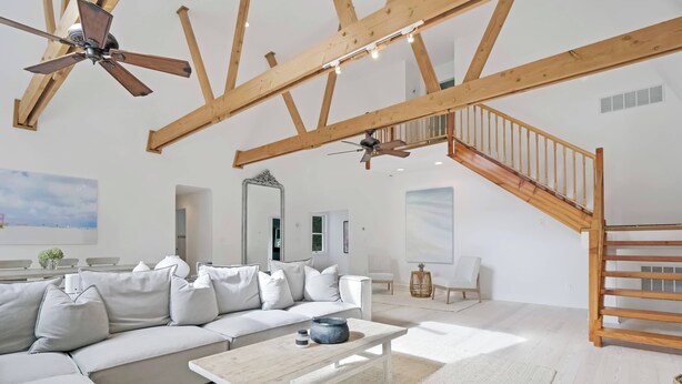 Ein geräumiges Wohnzimmer mit einem großen, weißen Ecksofa, einem Holztisch und einem Deckenventilator. Eine Holztreppe führt zu einer oberen Ebene.