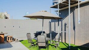 Un patio al aire libre con una mesa redonda, cuatro sillas, un paraguas grande, una parrilla y un taburete de madera.