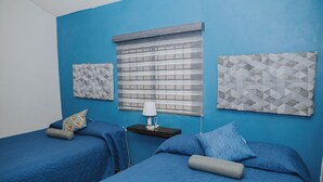 Una habitación con dos camas, cada una con ropa de cama y almohadas azules. Hay una pequeña mesa con una lámpara y un teléfono entre las camas. Las paredes están pintadas de azul, y hay tapices decorativos sobre las camas.