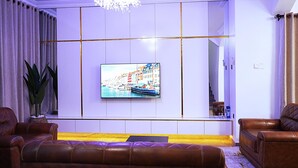 Ett modernt vardagsrum med en stor platt-TV som hänger på en vit vägg, lädersoffor, en gul matta och en stor krukväxt.