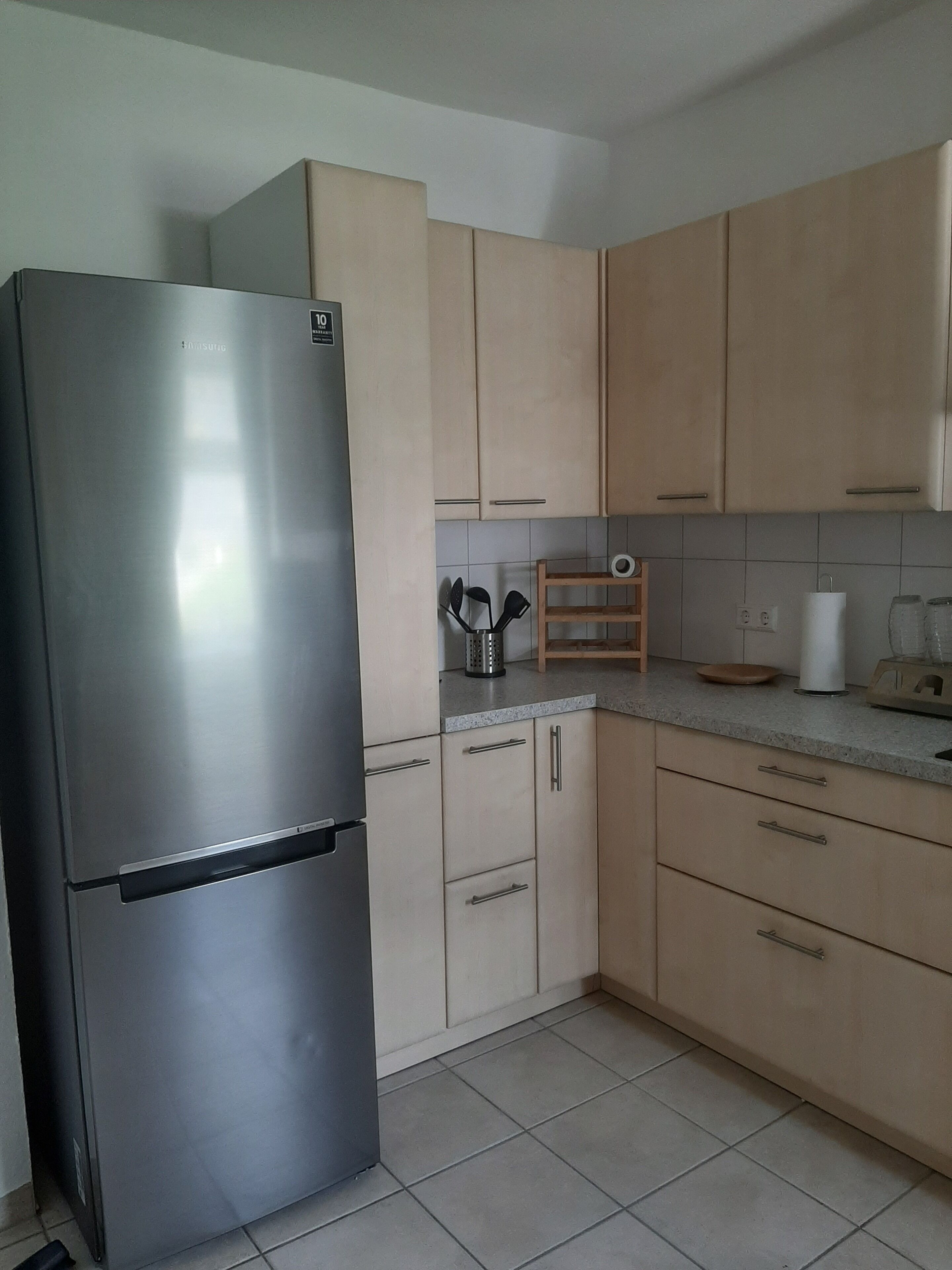 Uma cozinha com armários de madeira clara, uma geladeira de aço inoxidável e piso revestido de azulejos.