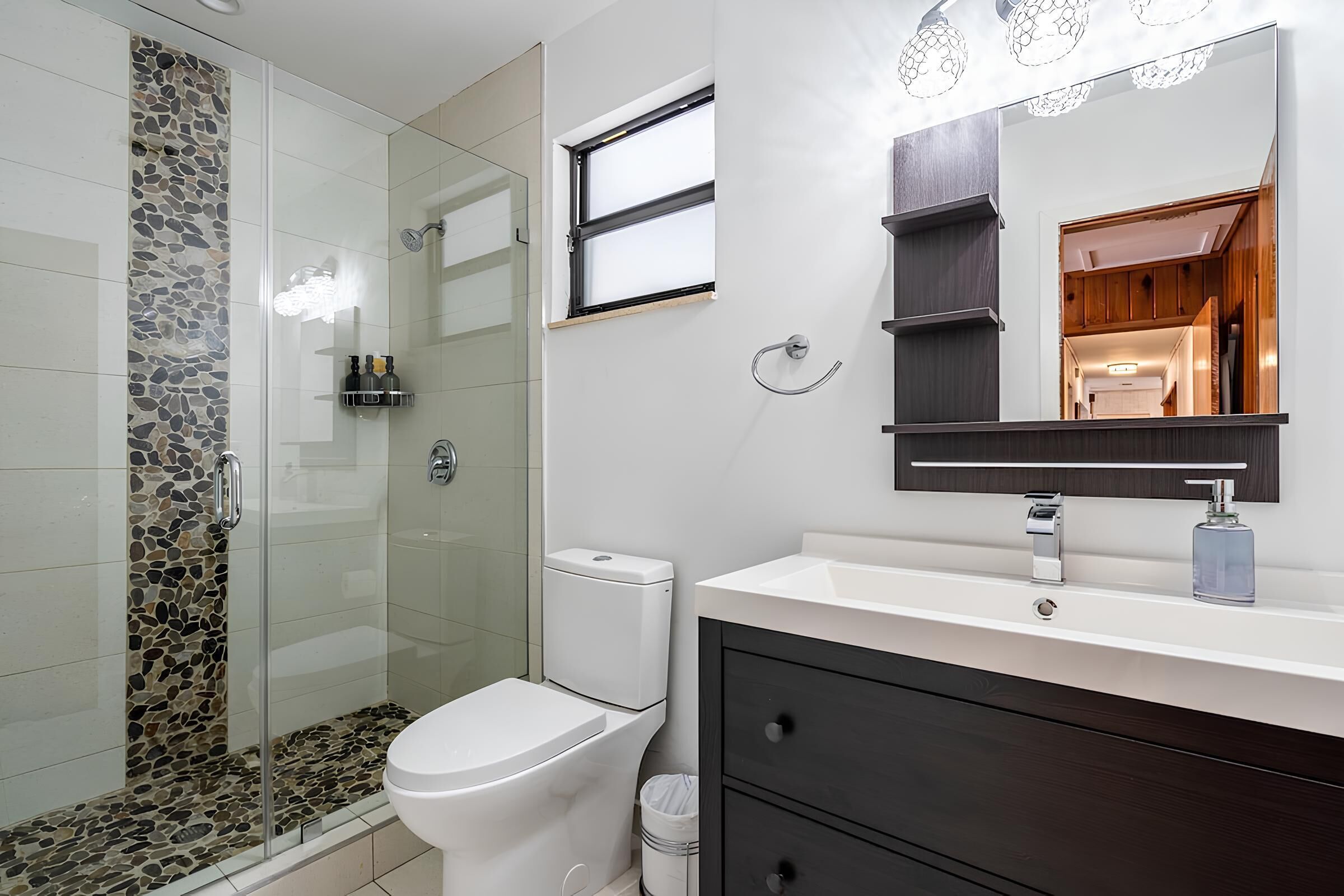 Une salle de bain moderne avec une douche à parois de verre, des toilettes blanches, un meuble-lavabo avec des placards sombres et un grand miroir.