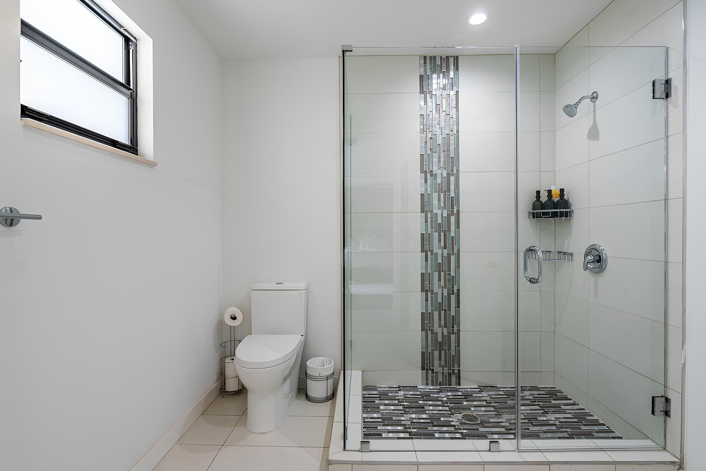 A modern bathroom with a glass shower enclosure, a toilet, and a small shelf with toiletries.