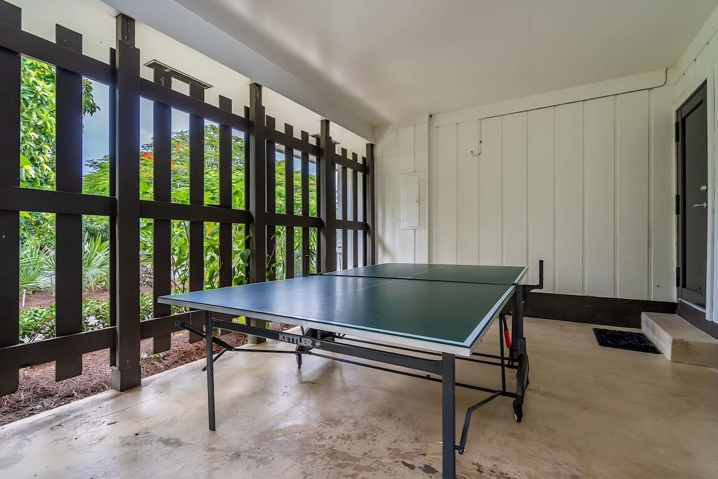 A ping pong table in a room with a concrete floor, white walls, and a wooden fence with greenery outside.
