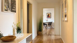 Un pasillo con piso de madera, una recepción con un cartel de 'Welcome', un comedor con una mesa y sillas, y obras de arte enmarcadas en las paredes.