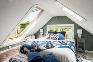 A bedroom with a sloped ceiling, skylight, bed with patterned bedding, bedside lamps, and a small nightstand. - White Mtn Hot Tub Retreat The Bit O’ Honey (Woodstock)