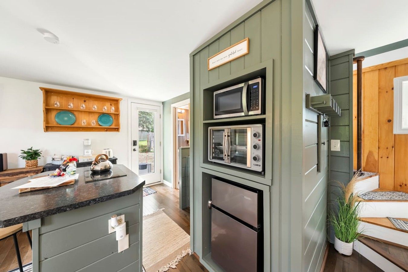 A kitchen with a built-in microwave, oven, and refrigerator, a wooden shelf with dishes, and a sign above the microwave.