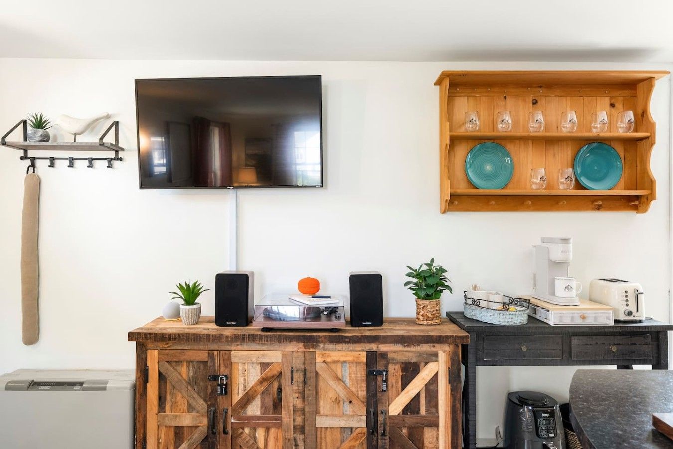 Ein modernes Wohnzimmer mit einem Wand-Fernseher, einem Holzregal mit Dekotellern, einer Kaffeemaschine und einer Stereoanlage.