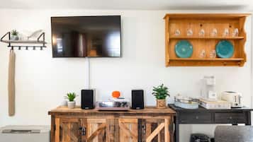 Uma sala de estar moderna com TV na parede, prateleira de madeira com pratos decorativos, cafeteira e sistema de som.