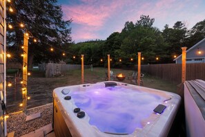 A hot tub with a wooden deck, surrounded by a wooden fence, string lights, and a fire pit.