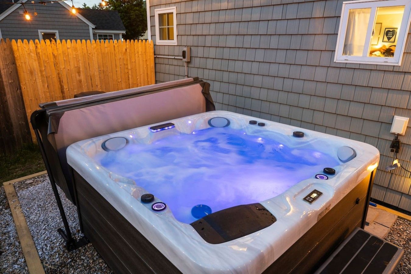 A hot tub with a wooden deck, a wooden fence, and a house in the background.