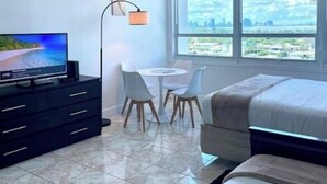 A modern hotel room with a bed, a TV, a desk with chairs, and large windows offering a city view. - Cute Studio Bay view! (Miami)