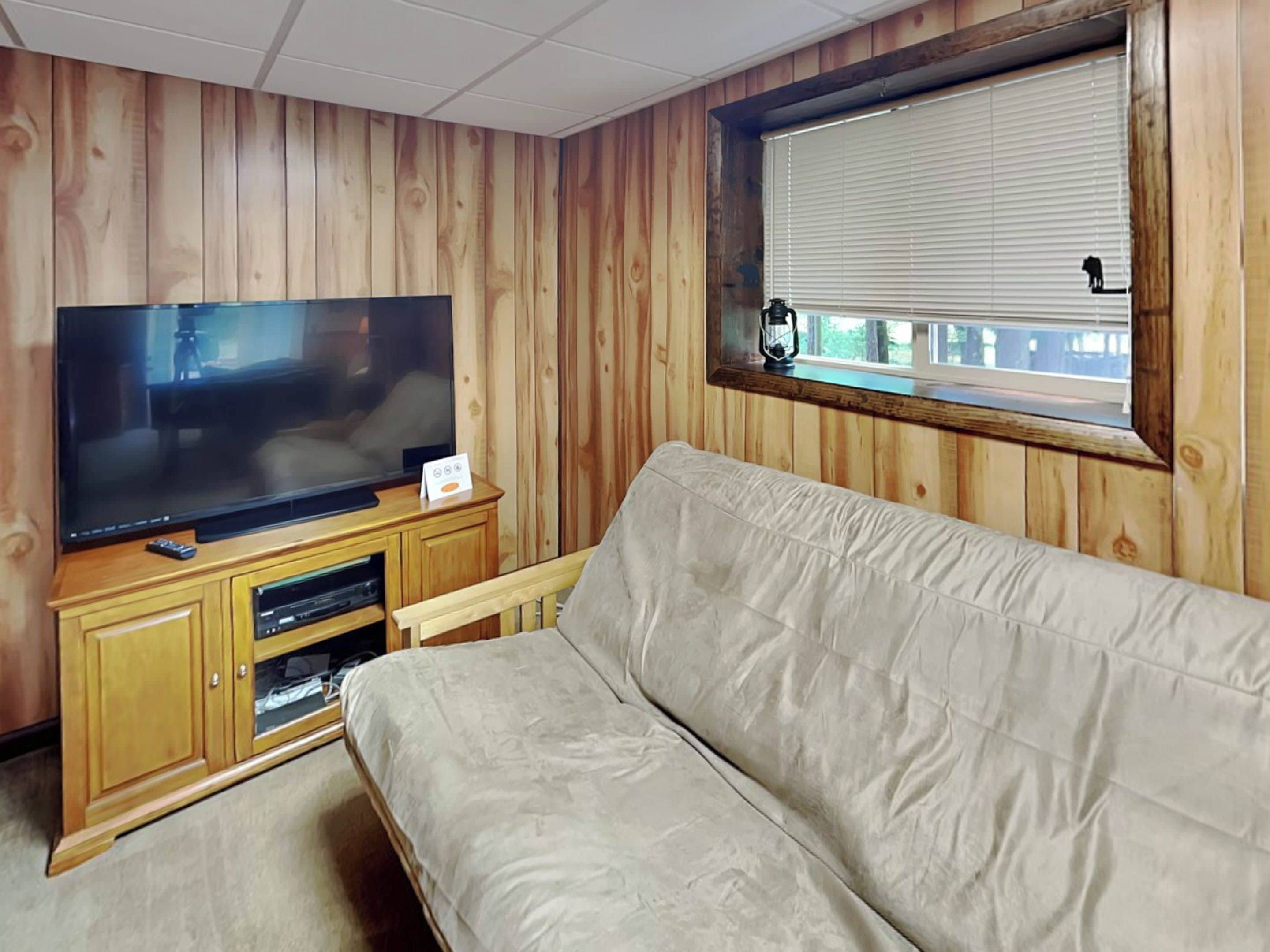 Una stanza con parete rivestita in legno, una grande TV a schermo piatto su un mobile in legno, un divano beige e una finestra con persiane.