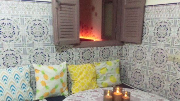 A cozy room with a bed, pillows, a table with candles, and a window with a view of a fire. - private room near taxi station and airport 
 (Inezgane)