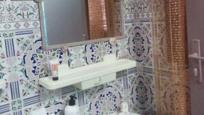 A bathroom with a white sink, a mirror above it, and a shelf with toiletries. The walls are tiled with a geometric pattern. - private room near taxi station and airport 
 (Inezgane)