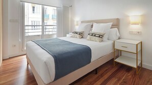 Moderni hotellihuone, jossa on suuri sänky, yöpöytä, lamppu ja ikkunasta avautuva näkymä kaupunkiin.
