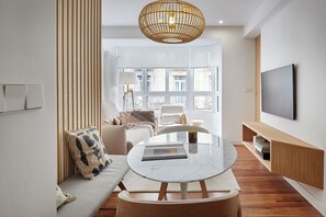 Un salon moderne avec une table ronde en marbre, un canapé agrémenté de coussins à motifs, un luminaire suspendu en osier et un grand téléviseur fixé au mur.