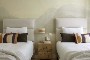 Une chambre d’hôtel avec deux lits, une table de chevet avec une lampe, des livres et une horloge.