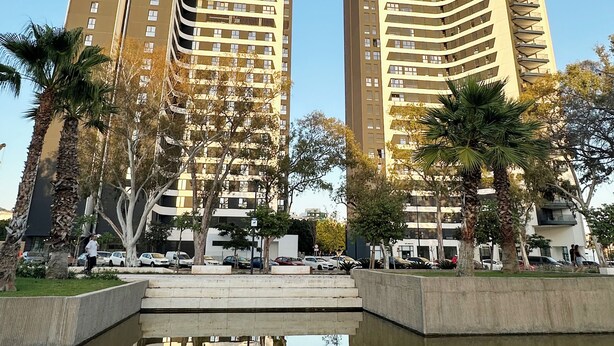 Dos edificis alts amb balcons, rodejats de palmeres i una petita piscina reflectant.