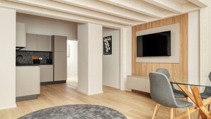 Una cocina moderna con armarios blancos y encimera negra, un comedor con sillas grises y mesa de vidrio, y un televisor montado en la pared.