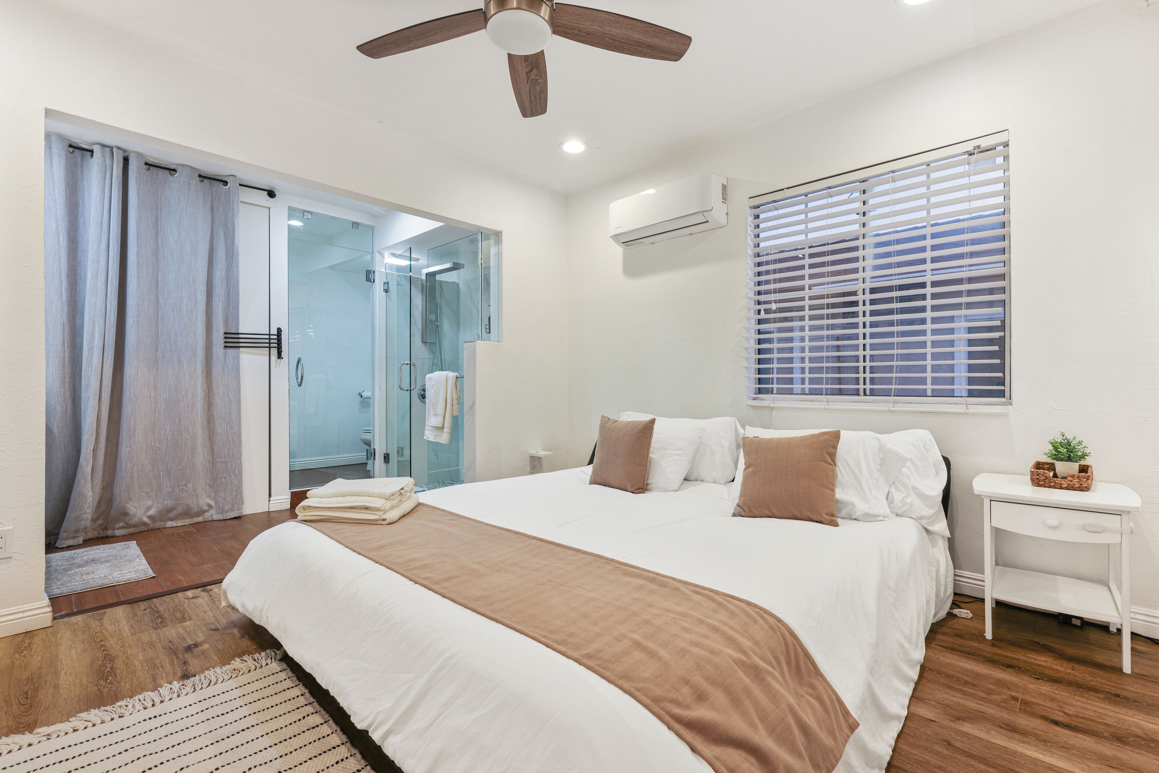 A bedroom with a large bed, a ceiling fan, a window with blinds, and a small bedside table with a plant.
