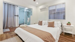 Ein Schlafzimmer mit einem großen Bett, einem Deckenventilator, einem Fenster mit Jalousien und einem kleinen Nachttisch mit einer Pflanze.