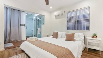 A bedroom with a large bed, a ceiling fan, a window with blinds, and a small bedside table with a plant.
