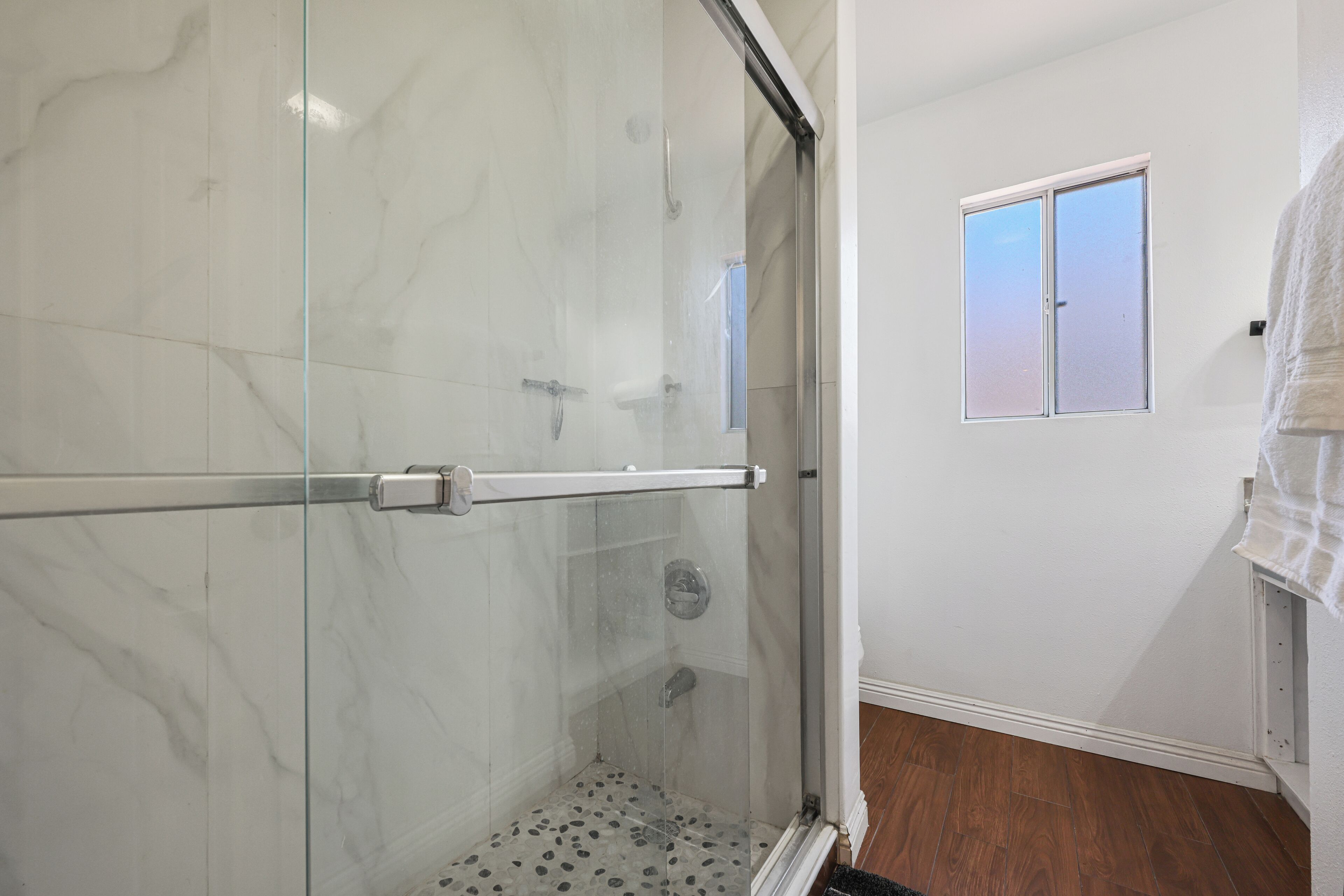 A bathroom with a glass shower enclosure, a small window, and a wooden floor.