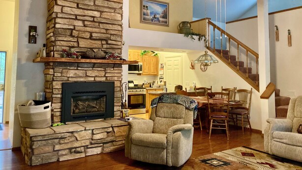 A spacious living room with a stone fireplace, comfortable seating, a dining area, and a staircase leading to an upper level. - Roomy Cabin in TALL Pines w/ Fenced Yd, A/C,  4  Bd/2 Ba, 75" TV + 593 movies (Pinetop)