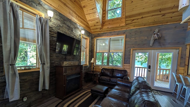 Interior acogedor de una cabaña con chimenea, sofá de cuero y grandes ventanales que ofrecen vistas del exterior.