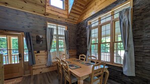 Una cabaña de madera con comedor que incluye mesa y sillas de madera, ventanas amplias con cortinas y una pared de piedra.