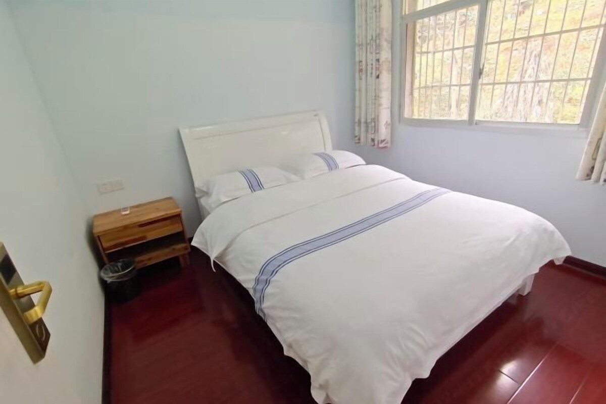 A bedroom with a bed, a wooden nightstand, a small trash bin, and a window with curtains.