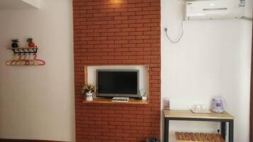 Een kamer met een bakstenen muur, een wandtelevisie, een klein tafeltje met een waterkoker en een plank met decoratieve objecten.