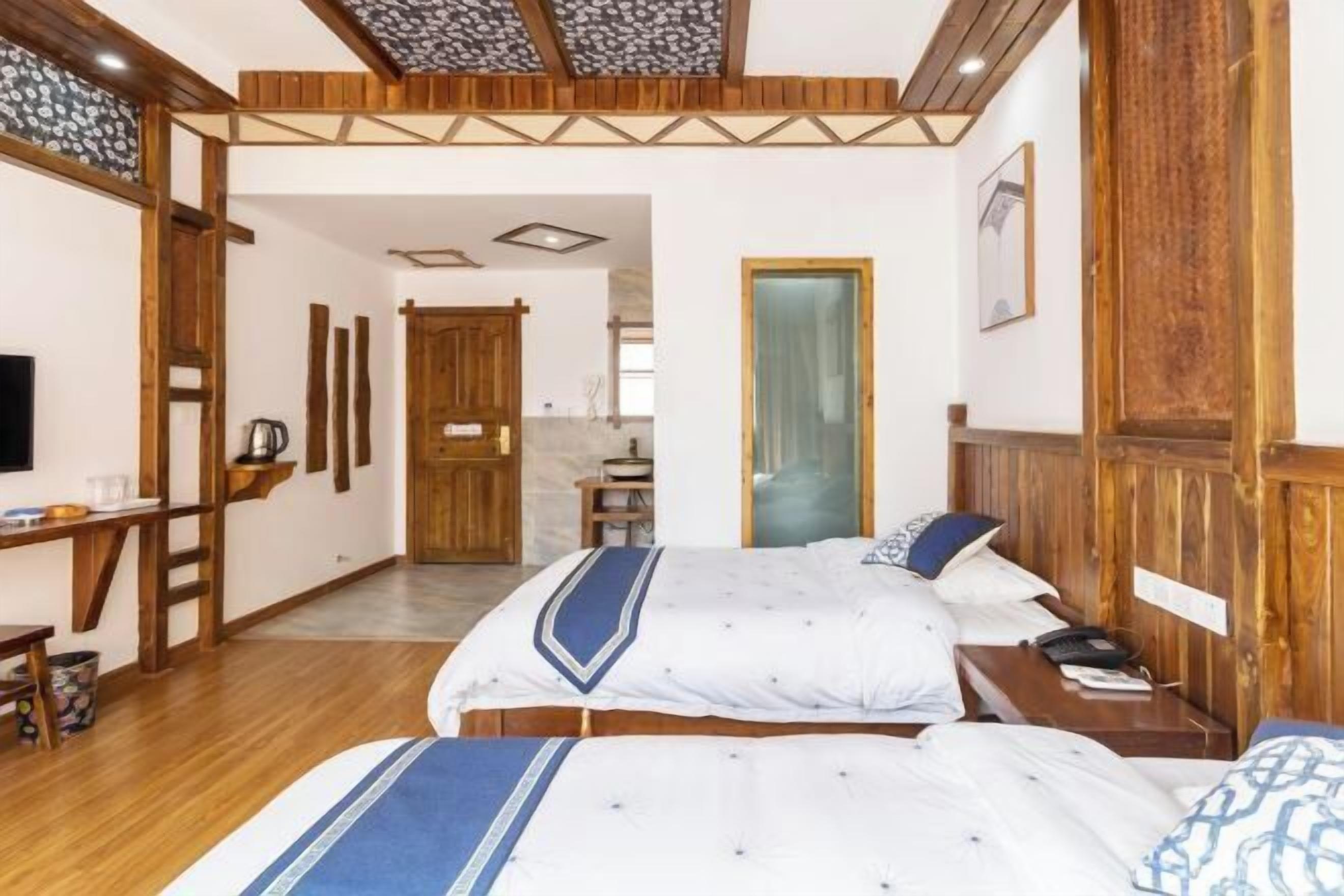Quarto de hotel com teto de madeira, cama com colcha azul e branca, mesa de cabeceira com abajur e guarda-roupa de madeira.