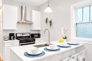 Una cocina moderna con armarios blancos, una campana extractora de acero inoxidable, una estufa a gas, un microondas y un lavabo con grifo. Hay individuales azules sobre la isla y una ventana con vidrio esmerilado.