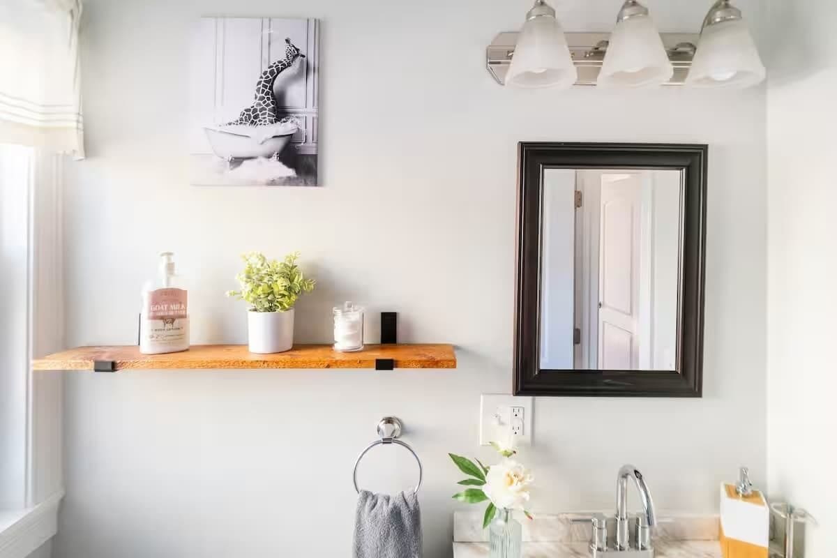 A bathroom with a wooden shelf holding toiletries, a framed mirror, and a wall-mounted light fixture.