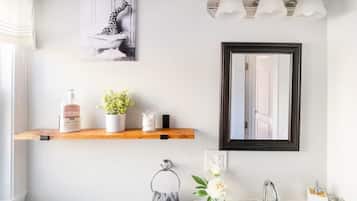 A bathroom with a wooden shelf holding toiletries, a framed mirror, and a wall-mounted light fixture.