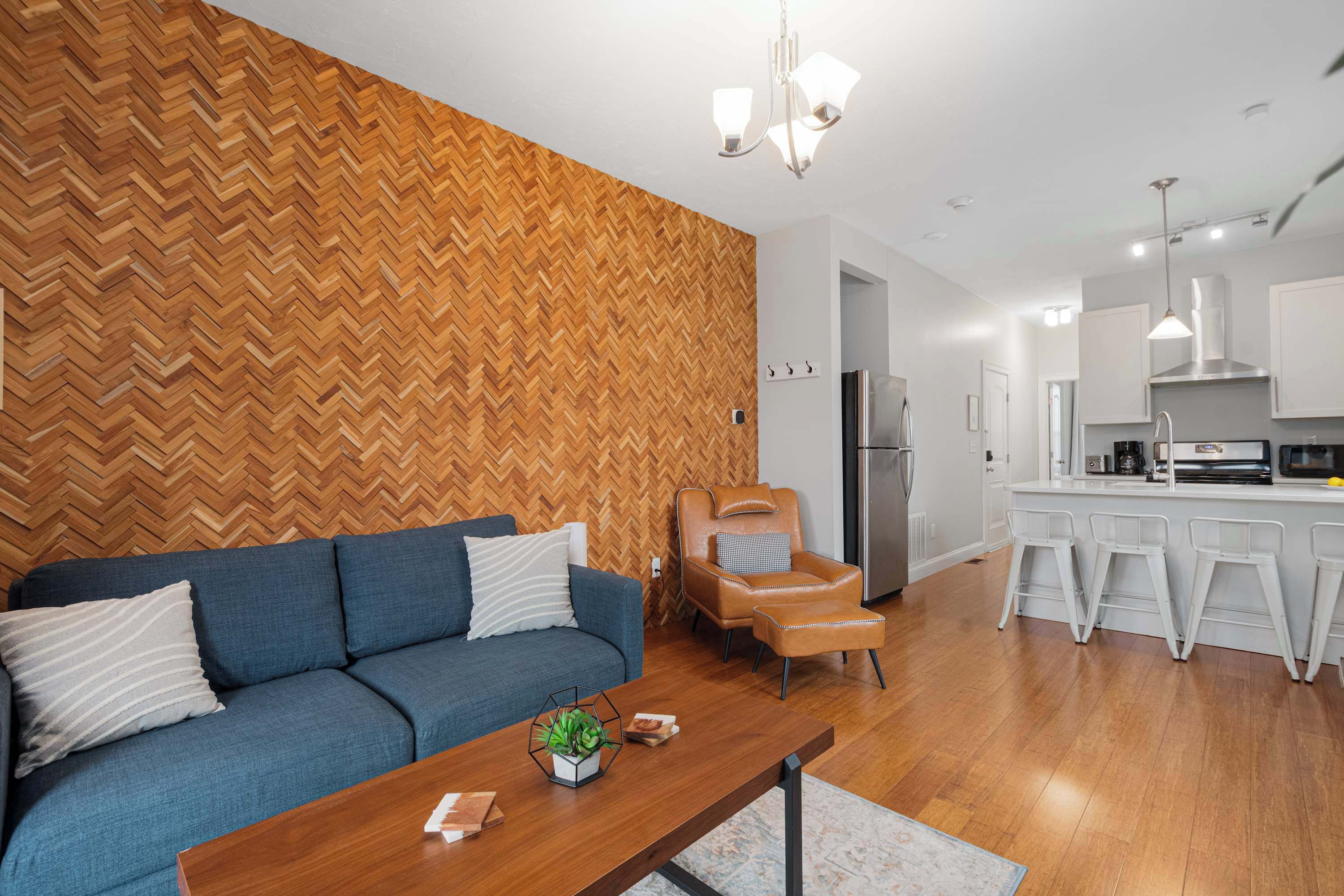 A modern living room with a herringbone wood wall, a blue sofa, a leather armchair, and a wooden coffee table with a plant.