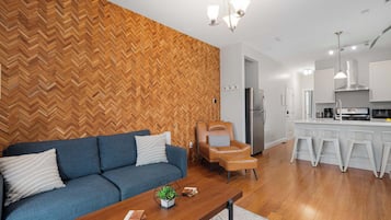 A modern living room with a herringbone wood wall, a blue sofa, a leather armchair, and a wooden coffee table with a plant.