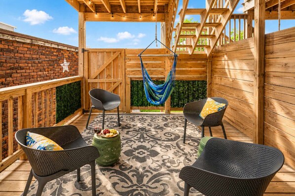 Una terraza de madera con dos sillas de mimbre negro, un taburete verde y una hamaca.