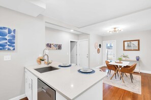 A modern kitchen with a central island, white cabinetry, and a dining area with a table and chairs. - Jennifer’s Chic Modern 1st Floor Apt Private Patio (Providence)