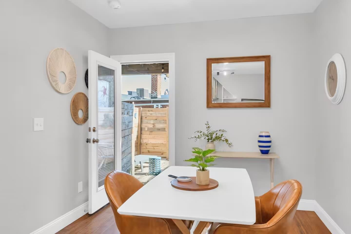 A dining area with a white table, two brown chairs, a potted plant, and a mirror on the wall.