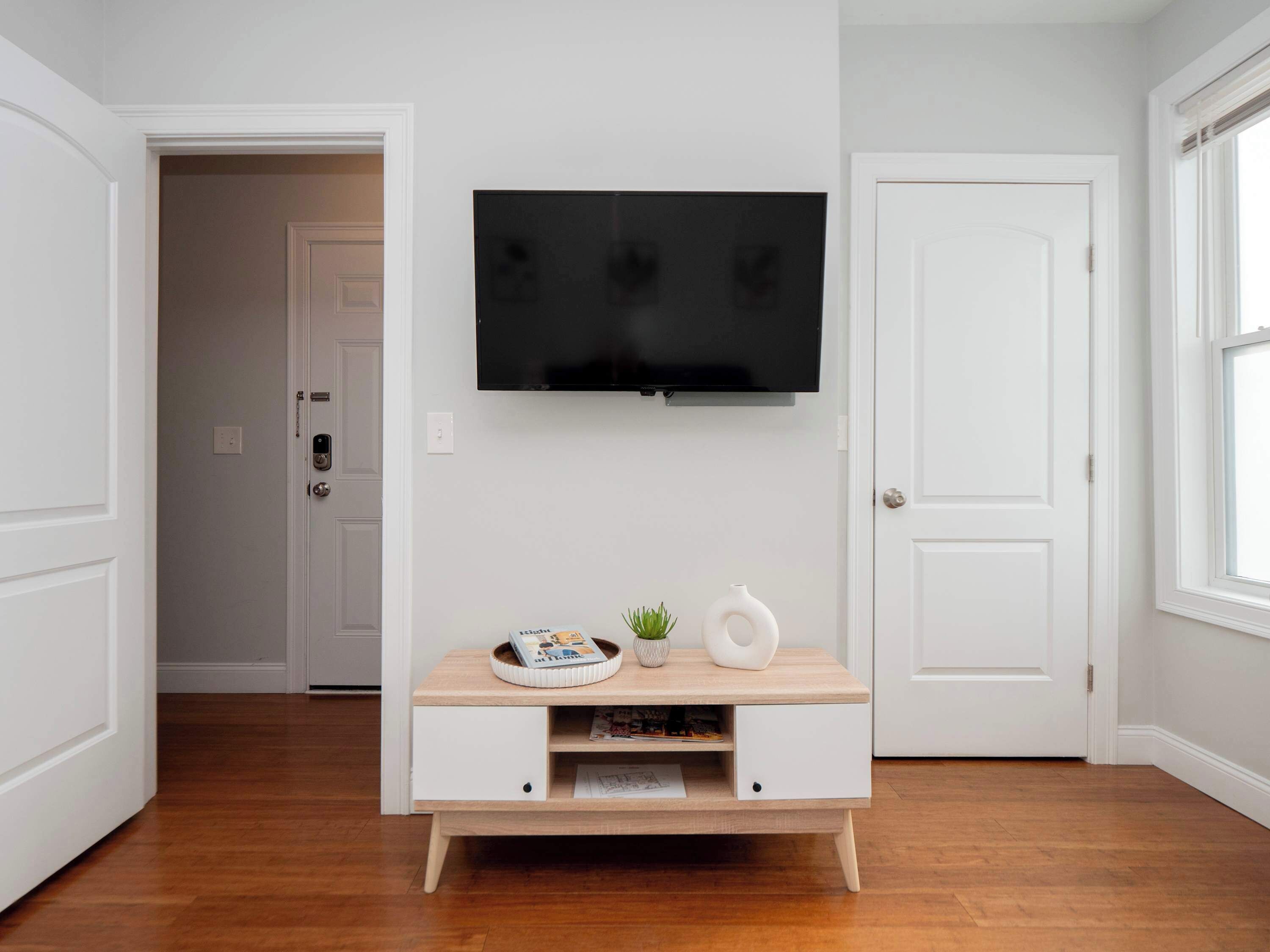 Een moderne woonkamer met een flatscreen televisie aan de muur, een houten console met decoratieve objecten, en een deur die naar een andere kamer leidt.