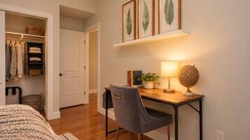 Un dormitorio con cama, escritorio y silla, estante con hojas enmarcadas y armario con ropa colgada.